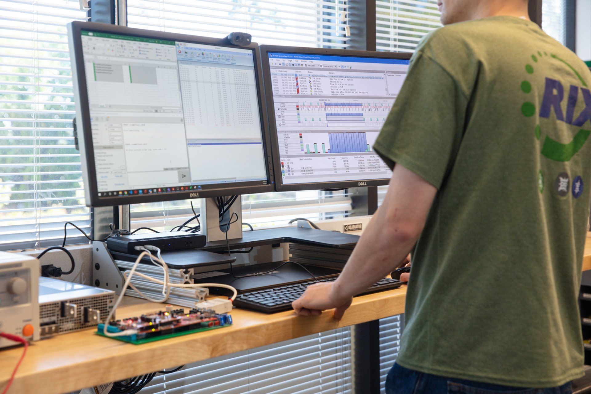 A man in a green shirt with the word rx on the back is standing in front of two computer monitors.