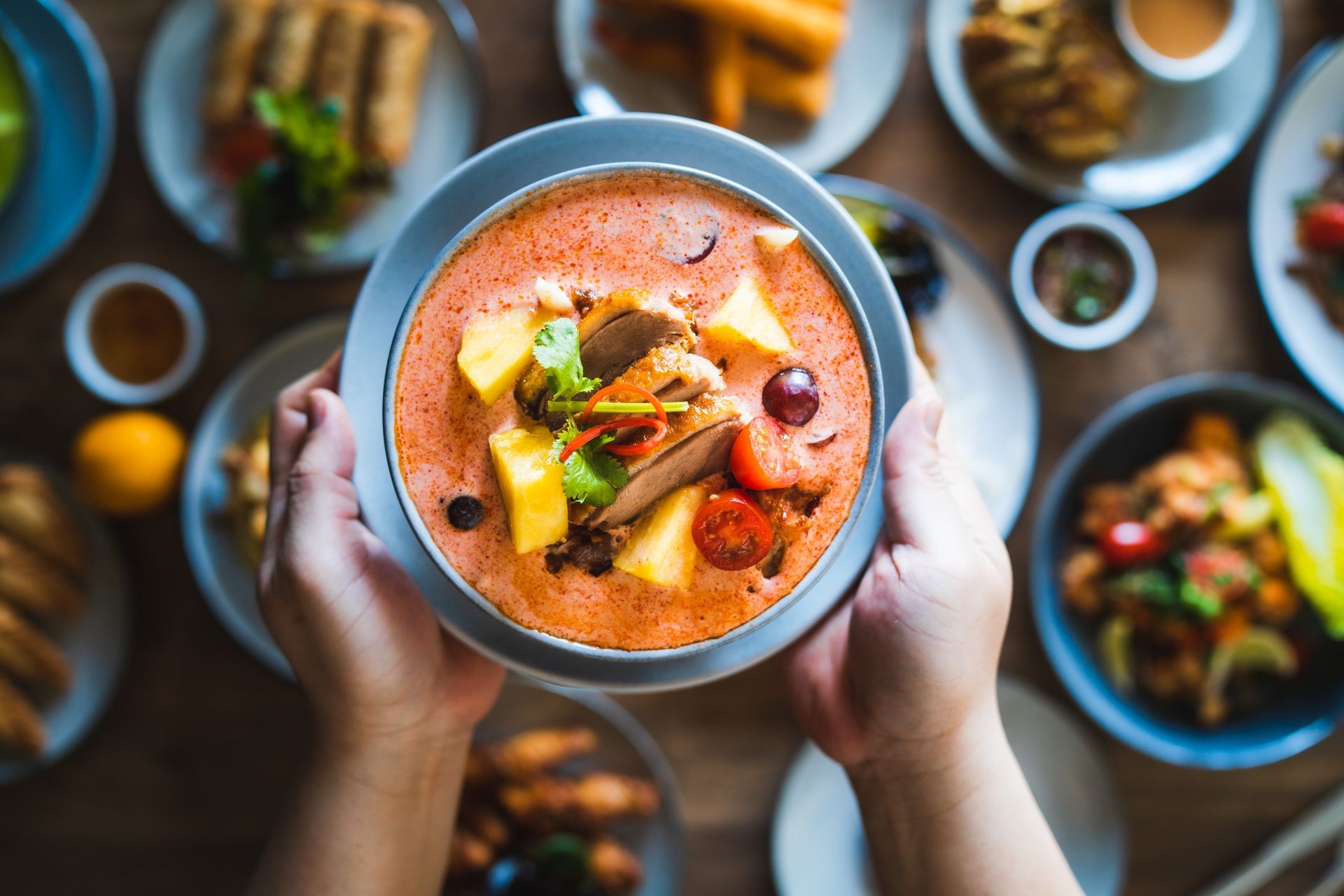 A person is holding a bowl of soup in front of a table full of food.