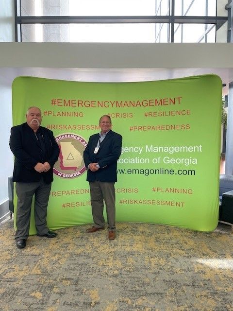 Two men are standing in front of a green sign that says emergency management.
