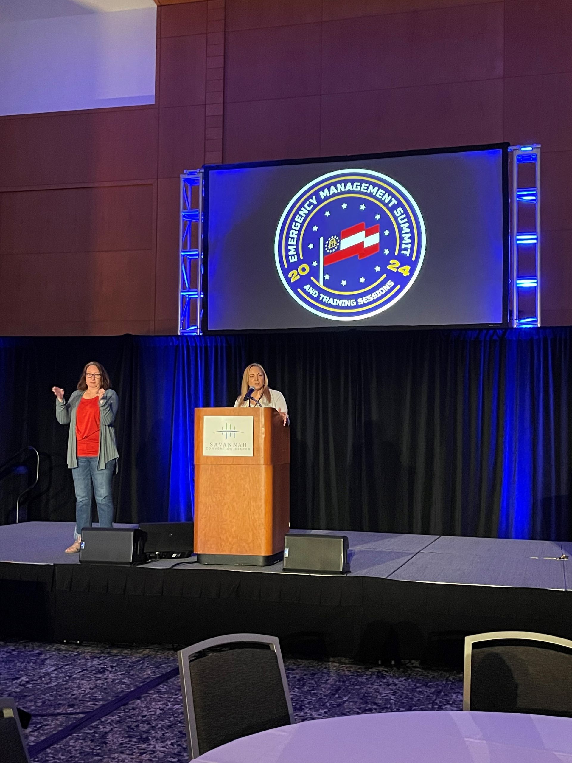 A woman stands at a podium in front of a large screen with a clock on it