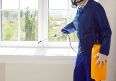 Pest control worker sprays around a window; wearing a respirator, blue jumpsuit, and yellow tank.