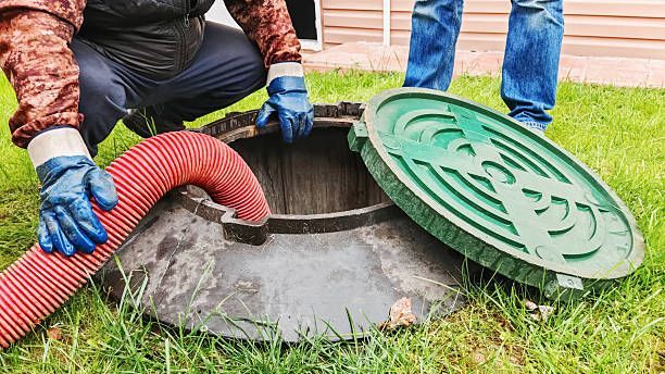 Man pumping out septic tank; hose inserted, lid open, gloves visible. Green lid, red hose.