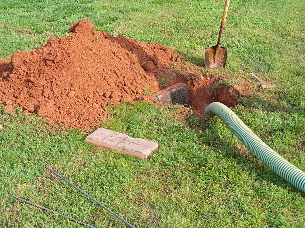Hole dug in grass with pile of red dirt, brick, shovel, and green hose.