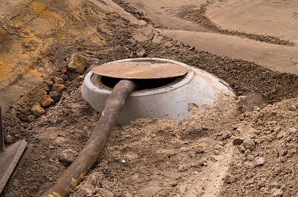 Concrete sewer access with metal lid and attached pipe, surrounded by dirt.