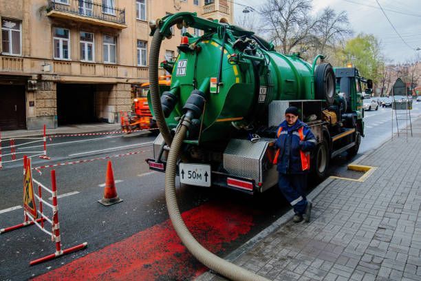 Green sanitation truck parked on a wet street with a worker. The hose extends onto the street, near red painted pavement.