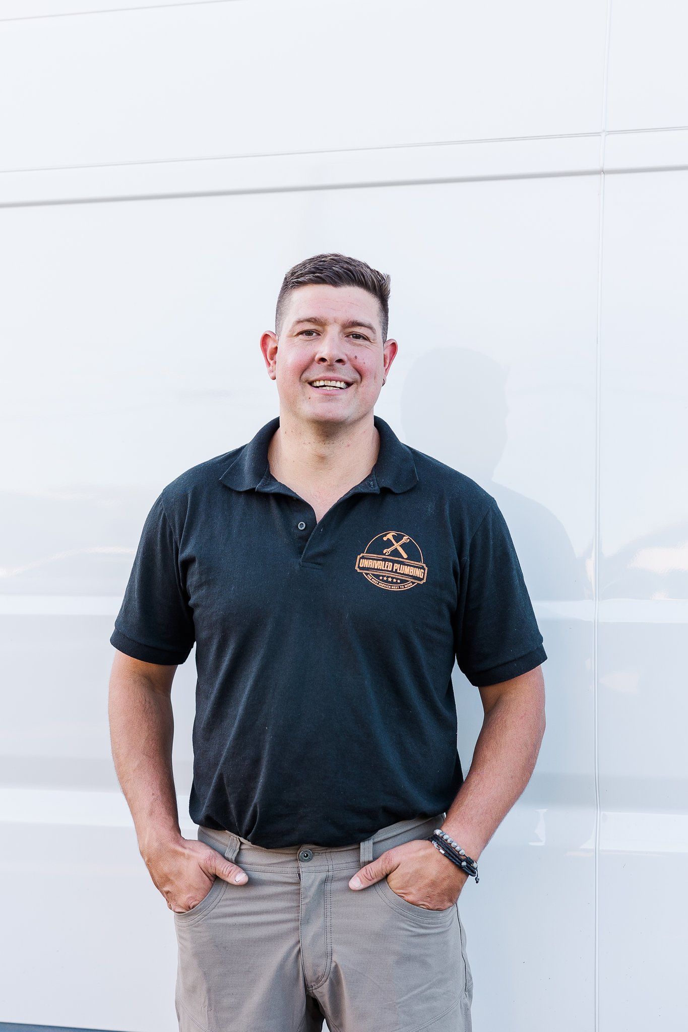 Man in black polo shirt and khakis, smiles, hands in pockets, stands against a white wall.