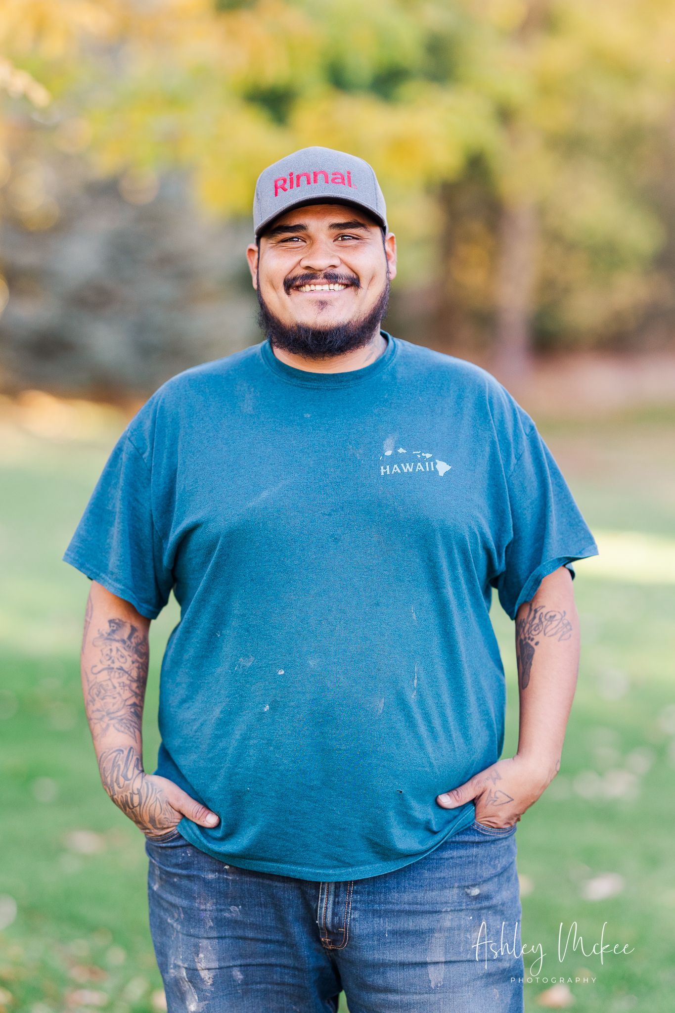Man with beard and hat smiling outdoors, hands in pockets. Wearing blue shirt and jeans. Man with beard and hat smiling outdoors, hands in pockets. Wearing blue shirt and jeans.