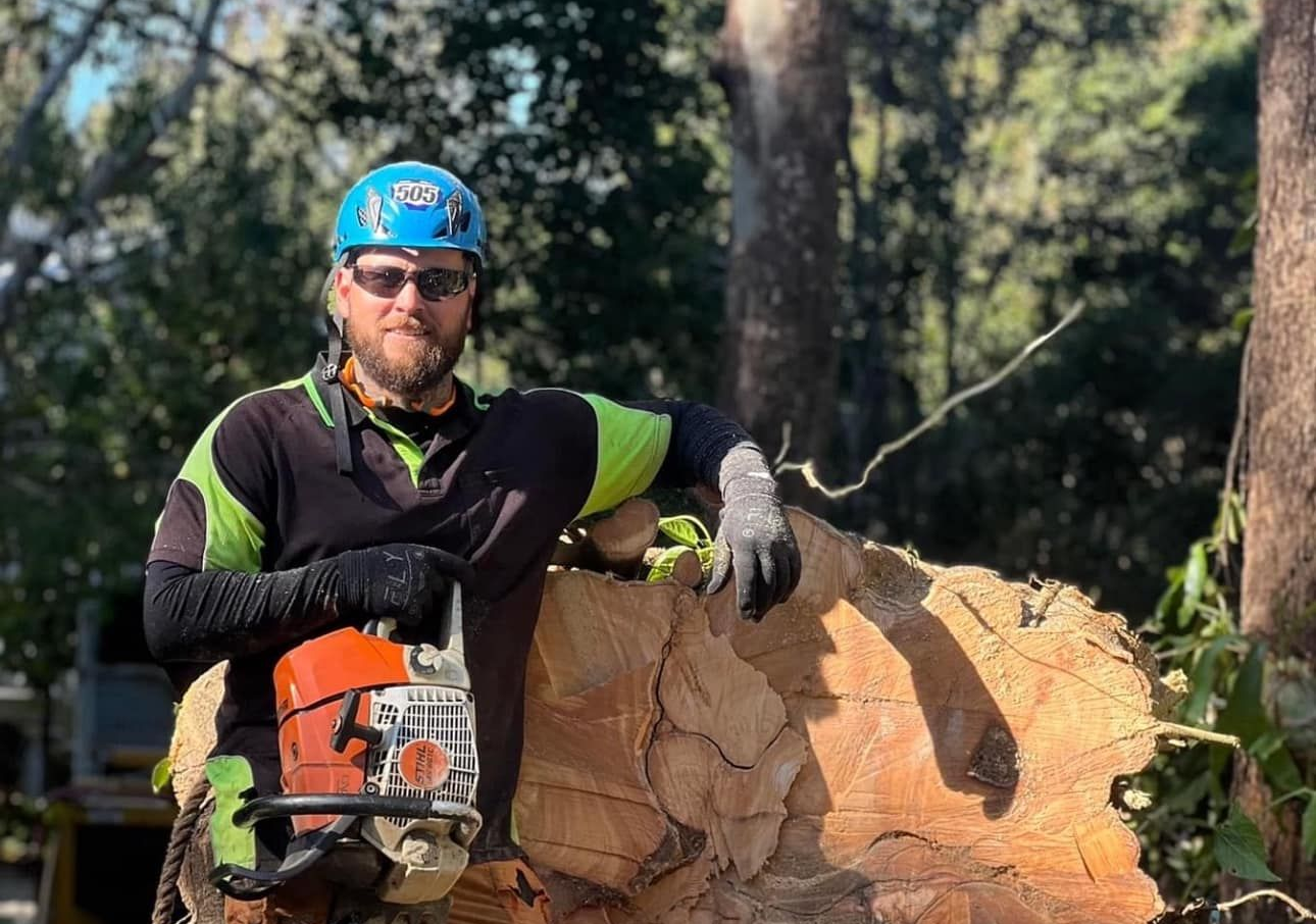 Arborist in safety gear leans on a large cut tree trunk, holding a chainsaw in an outdoor setting. Frisco Texas