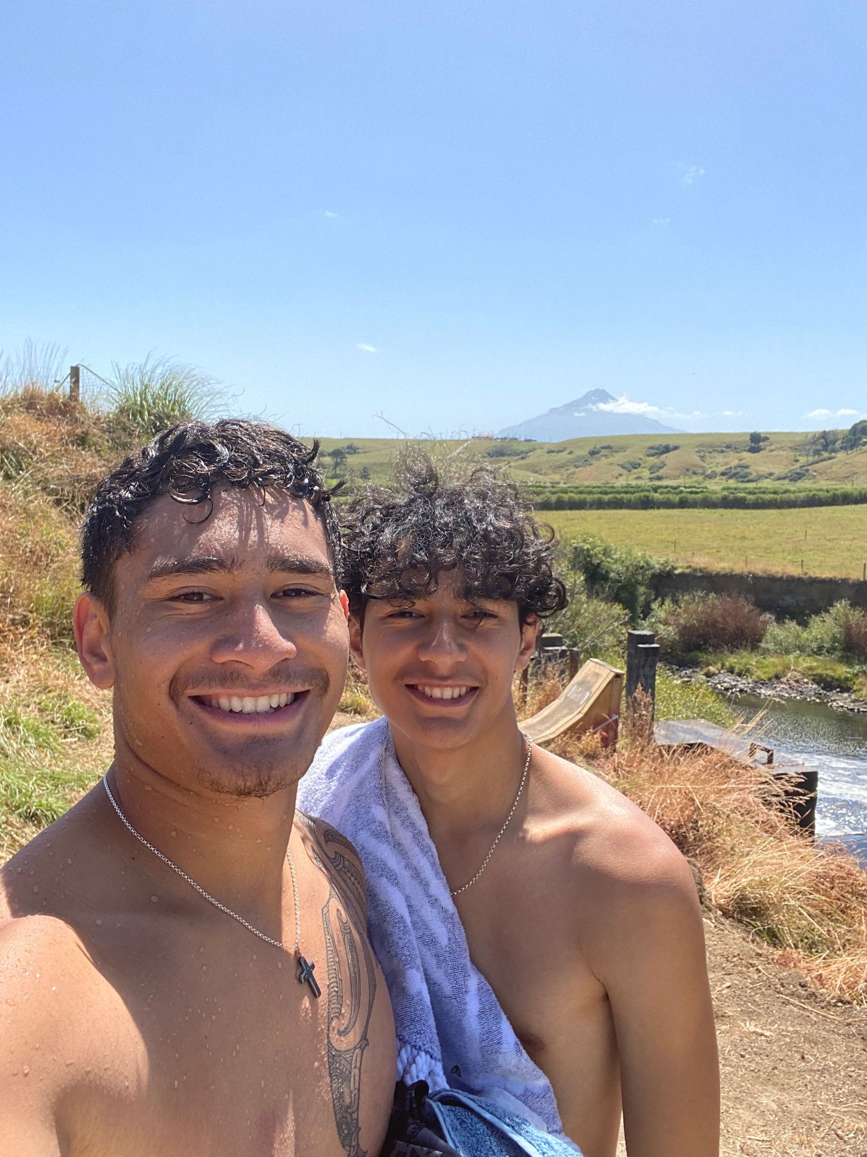 Two smiling young Māori men outdoors, near a stream, with mount Taranaki in the background. Sunny day.