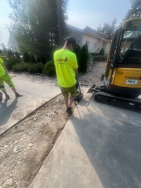 Rakennustyöntekijä tiivistää soraa tärylevyllä jalkakäytävällä kaivinkoneen vieressä.