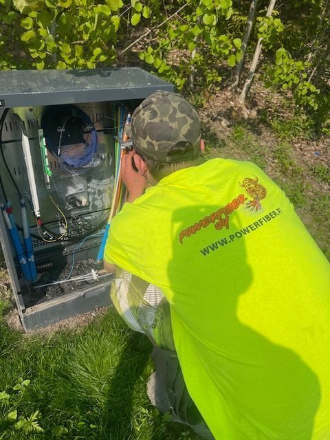 Neonkeltaiseen paitaan pukeutunut henkilö työskentelee harmaan sähkökeskuksen parissa ulkona kasvillisuuden lähellä.