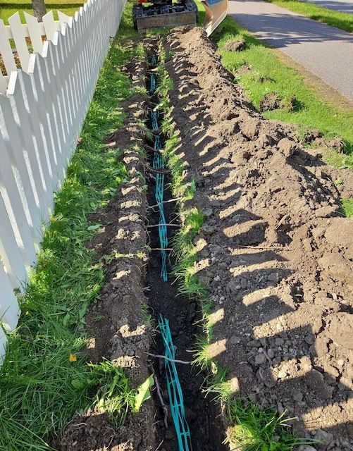 Valkoisen lauta-aidan viereen kaivettu oja, joka on täytetty vihreillä putkilla. Taustalla kaivinkone.