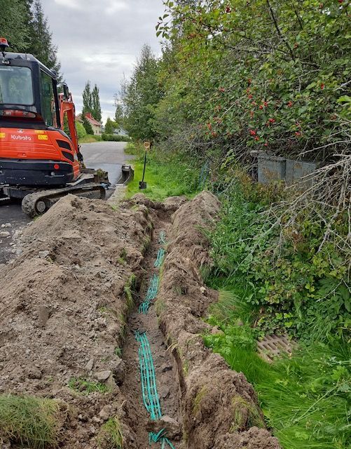 Kaivinkone vihreitä putkia sisältävän ojan vieressä tien laidassa, jossa on puita ja kasvillisuutta.