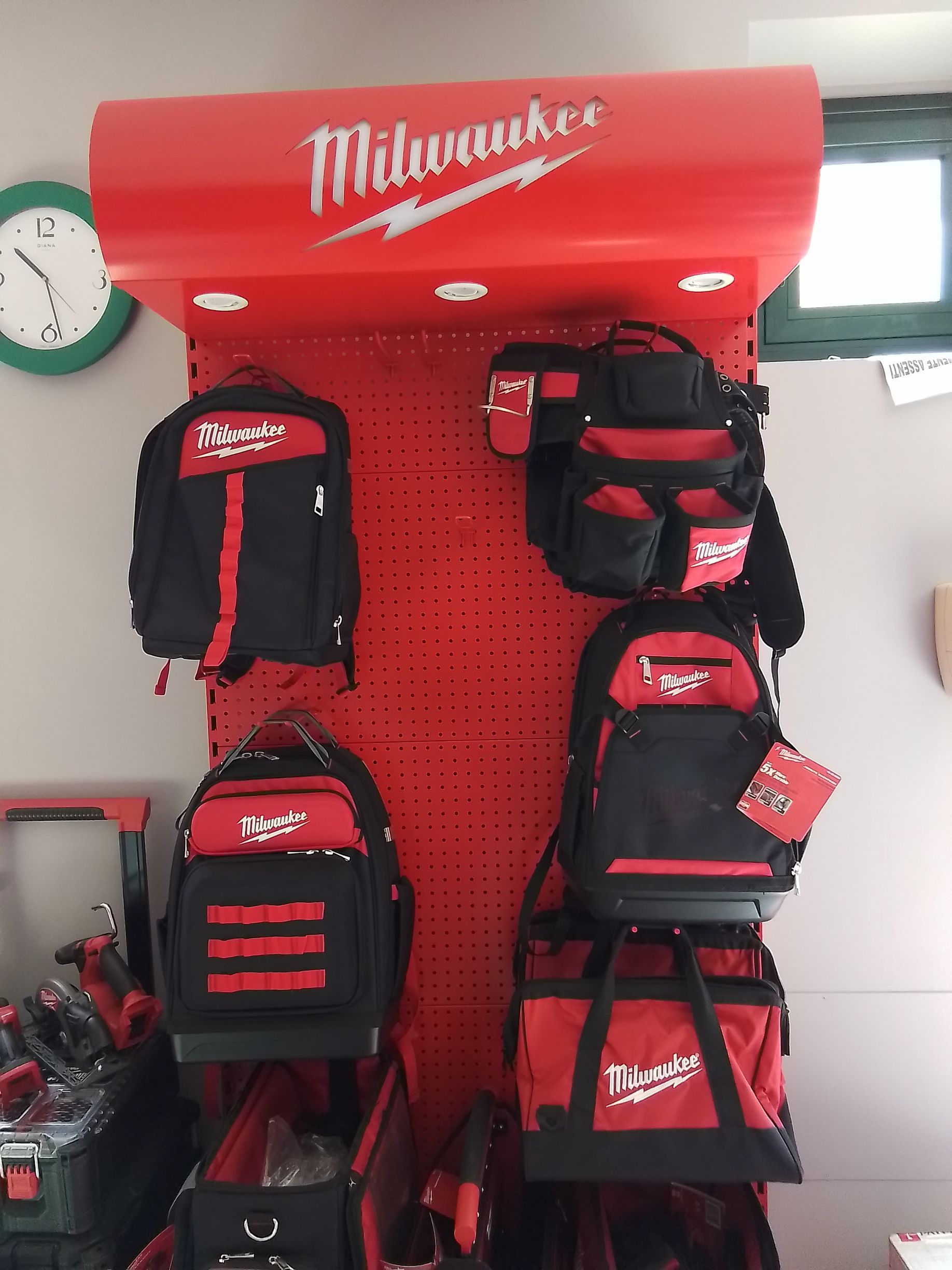 Borse portautensili e zaini rossi Milwaukee esposti su un pannello forato rosso, sotto un cartello Milwaukee.