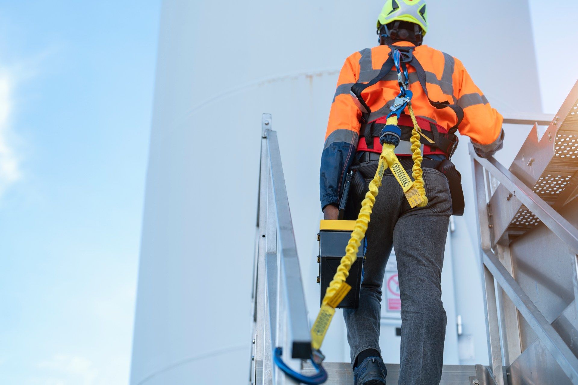 Un operaio sale su una scala di una turbina eolica, indossando un'imbracatura di sicurezza e un giubbotto arancione brillante contro il cielo azzurro.