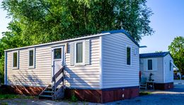 Two white mobile homes with red trim and gray roofs, surrounded by trees.