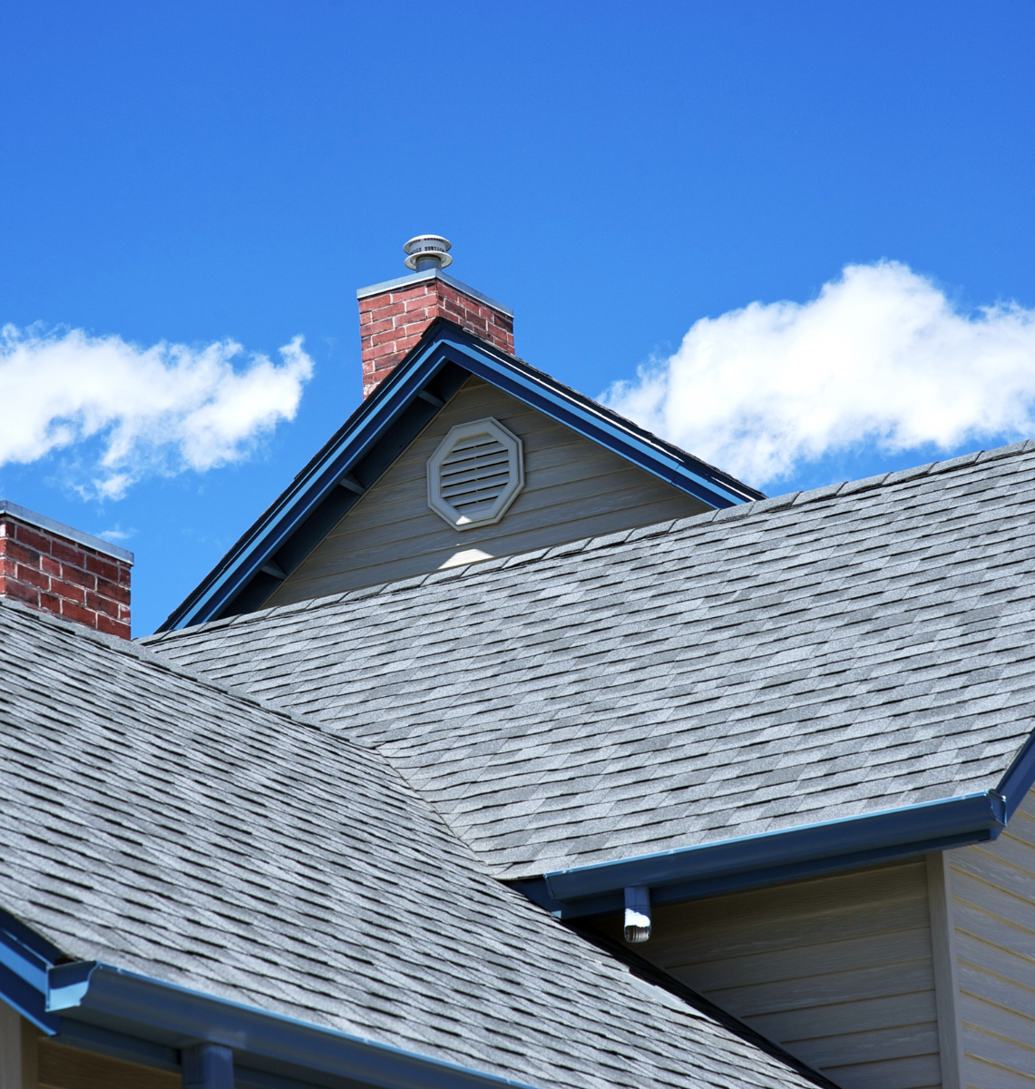 House rooftops with grey shingles, chimneys, blue trim against a blue sky with clouds.