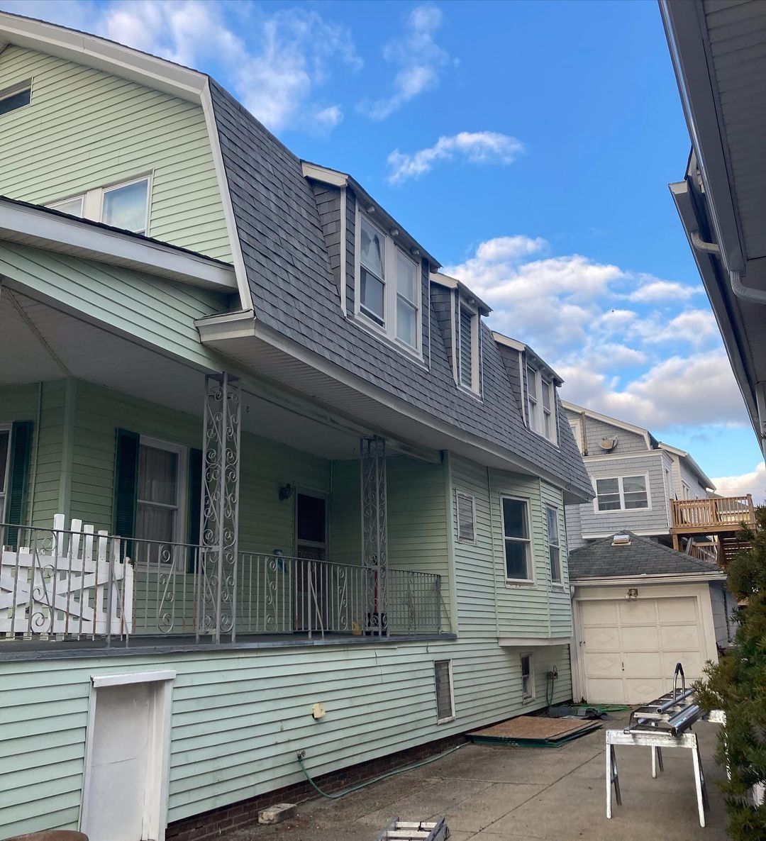 Light green house with gray mansard roof and a porch. Sunny day.
