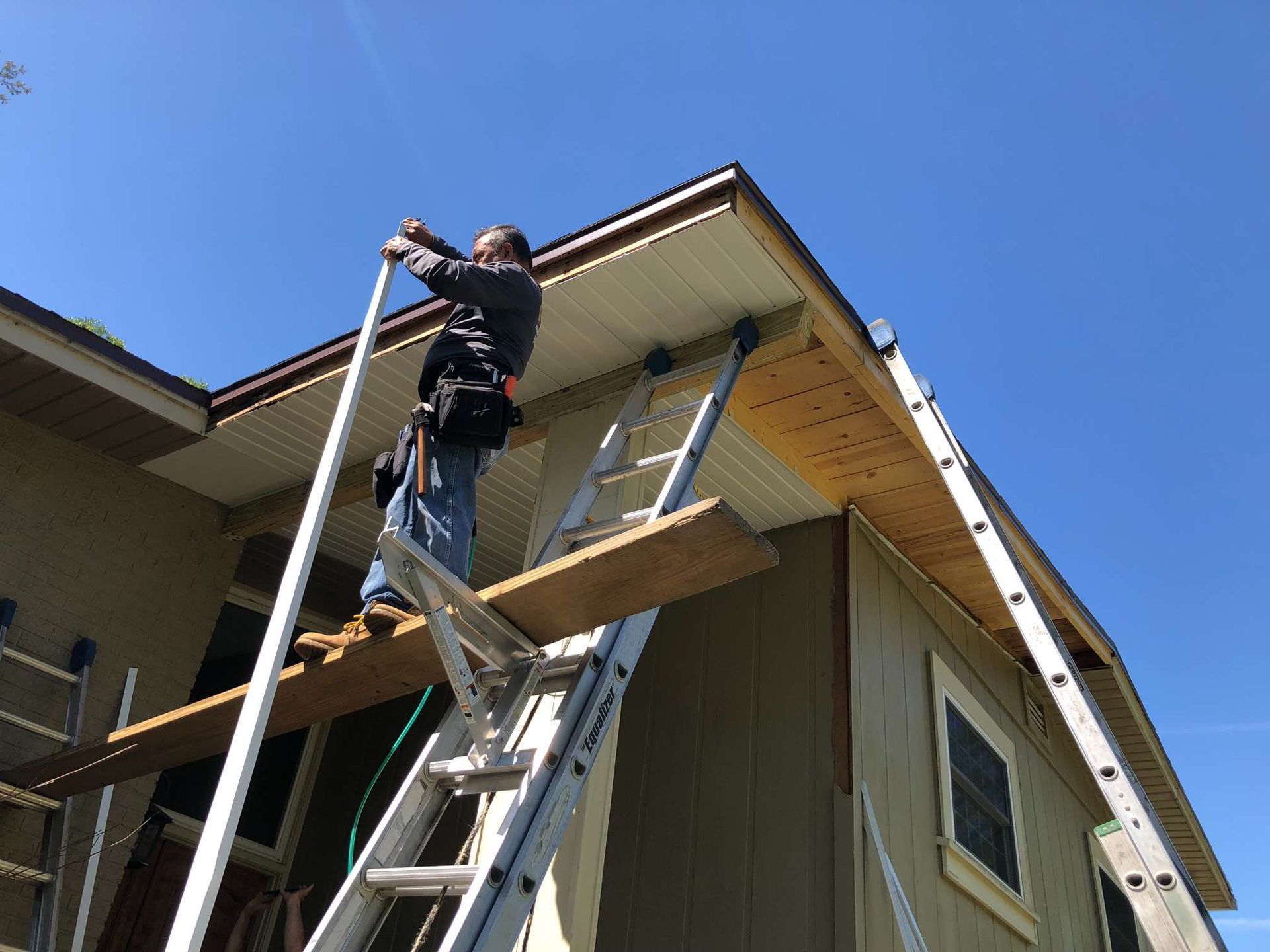 Man on a ladder installing soffit panels on a house exterior; sunny, blue sky.