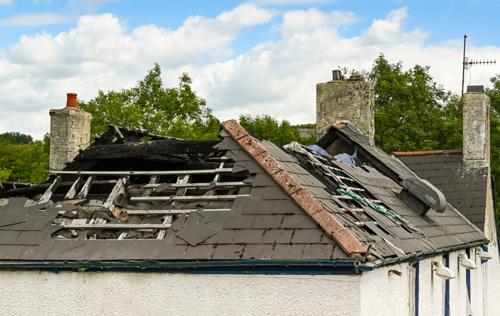 Damaged house roof with missing shingles and exposed beams, chimneys, and green trees in the background.