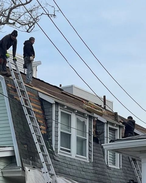 Three roofers working on a house. One on a ladder, two on the roof. Gray and white colors, with power lines overhead.