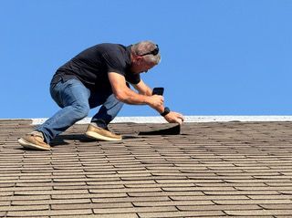 Workers replacing roof shingles on a two-story white house, ladder propped up.