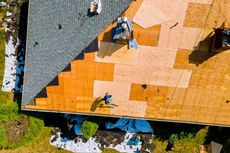 Aerial view of a roof partially re-shingled; a worker stands on the roof.