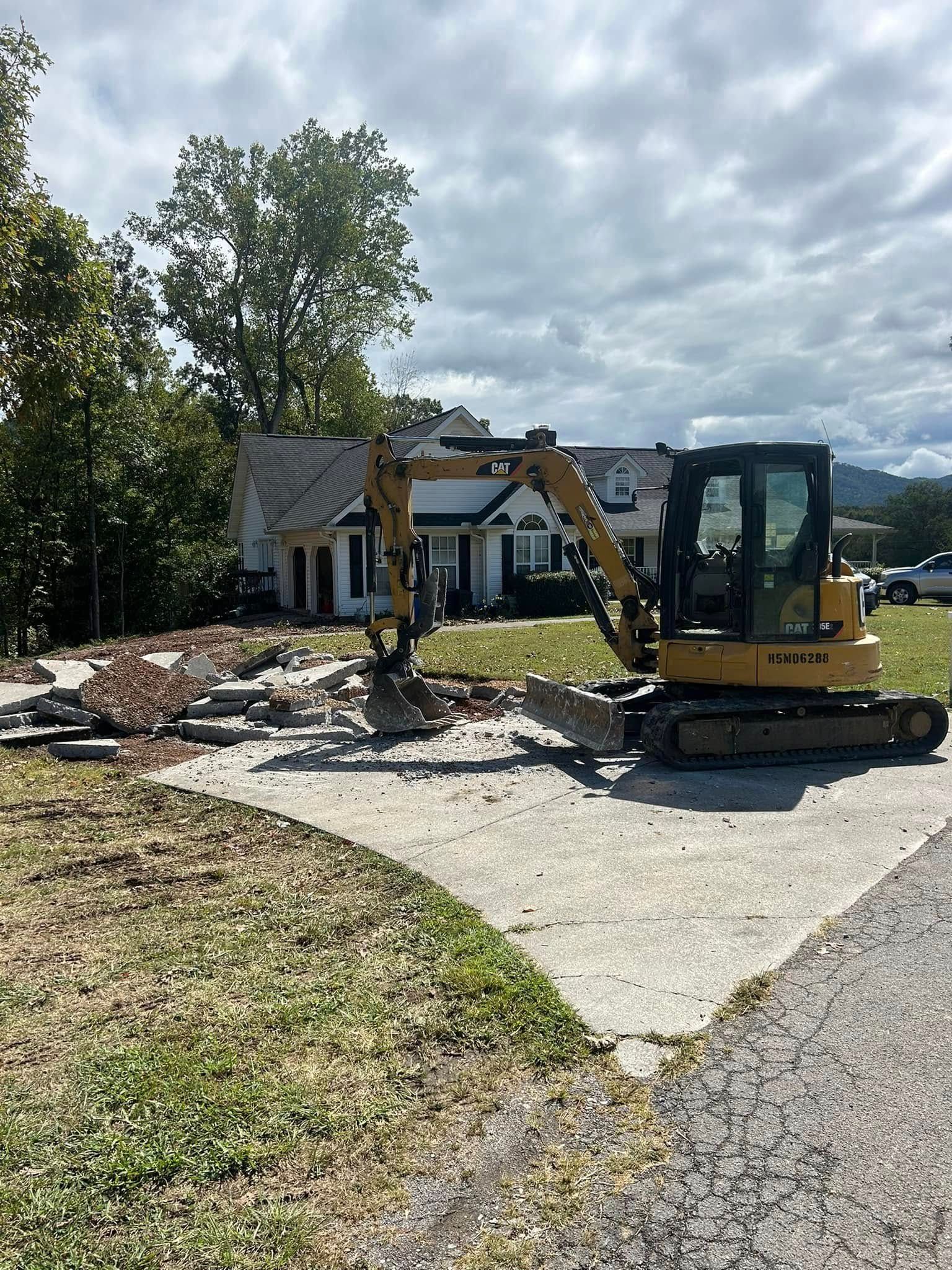 Yellow excavator demolishing a brick walkway in front of a white house on a cloudy day.