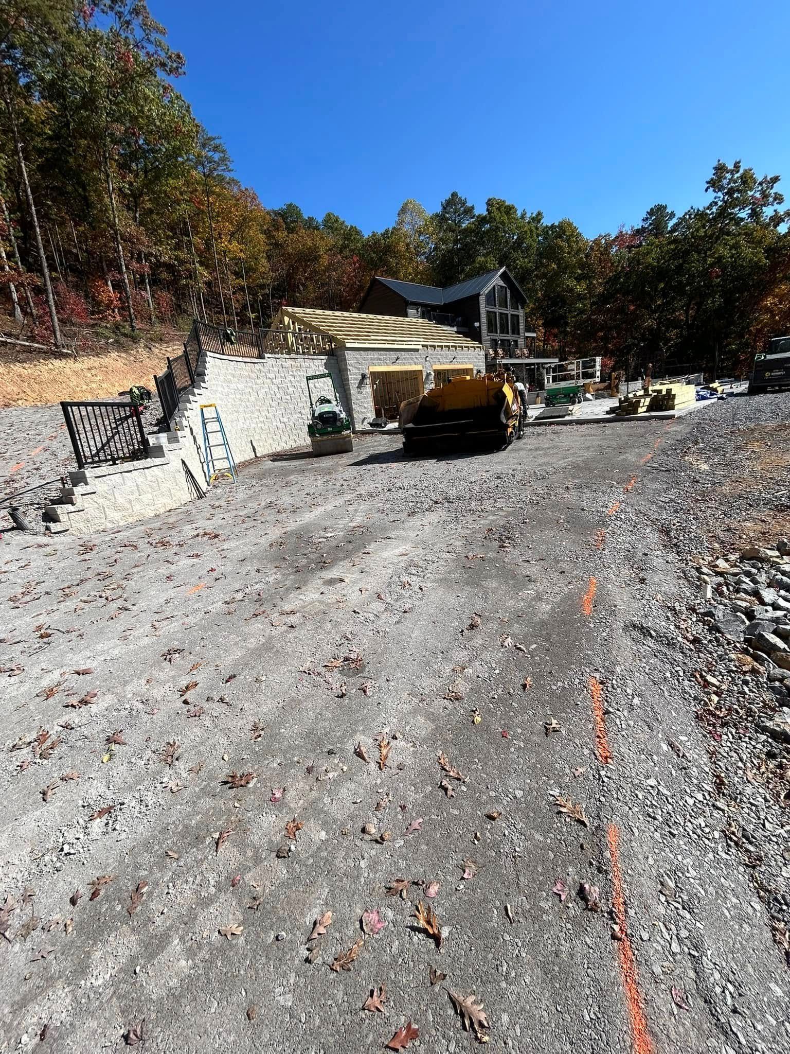 Construction site: gravel road leads to building platform with machinery under a blue sky, trees in the background.