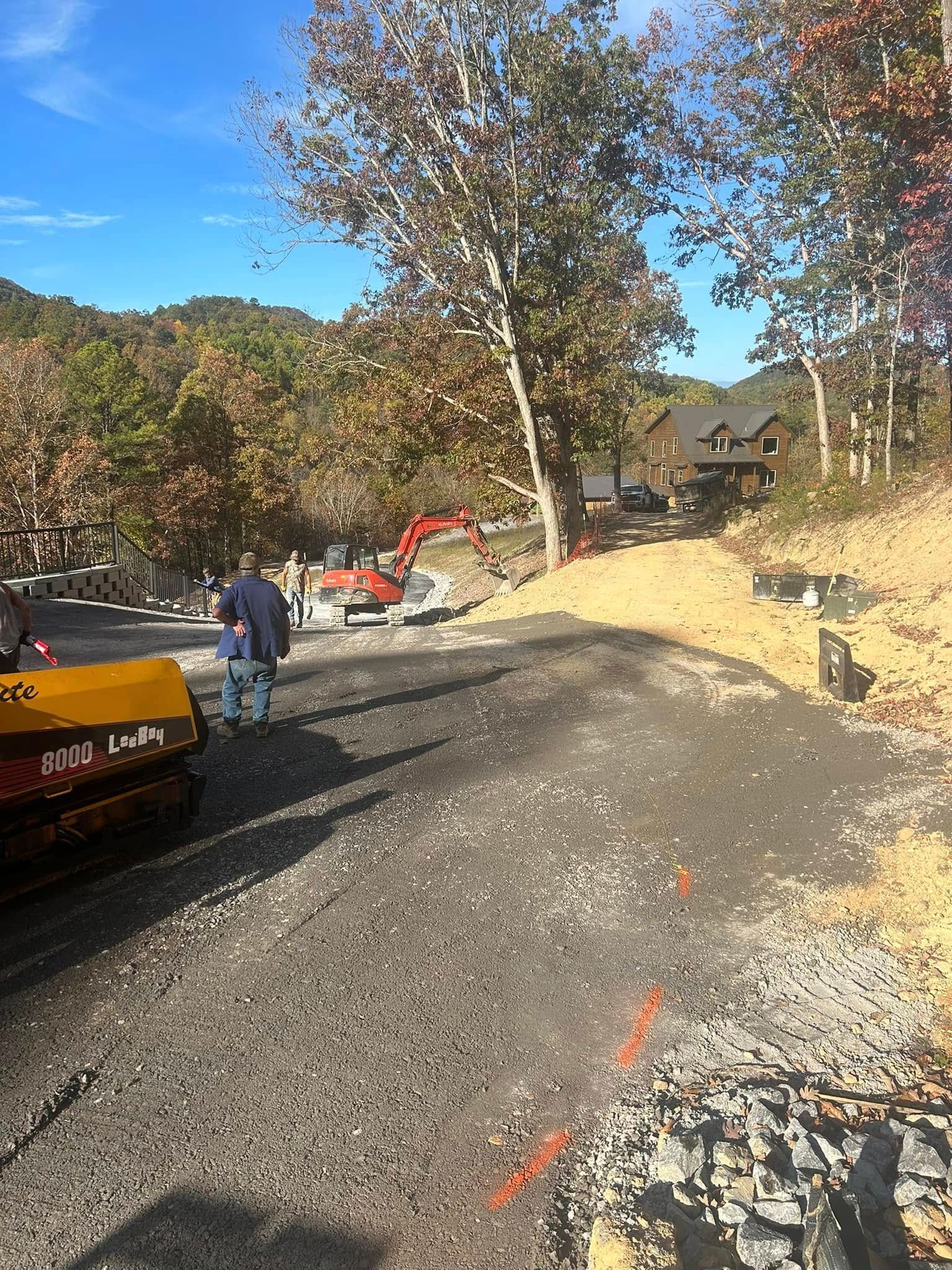 Road construction: asphalt being laid with machinery, worker nearby, trees in background.