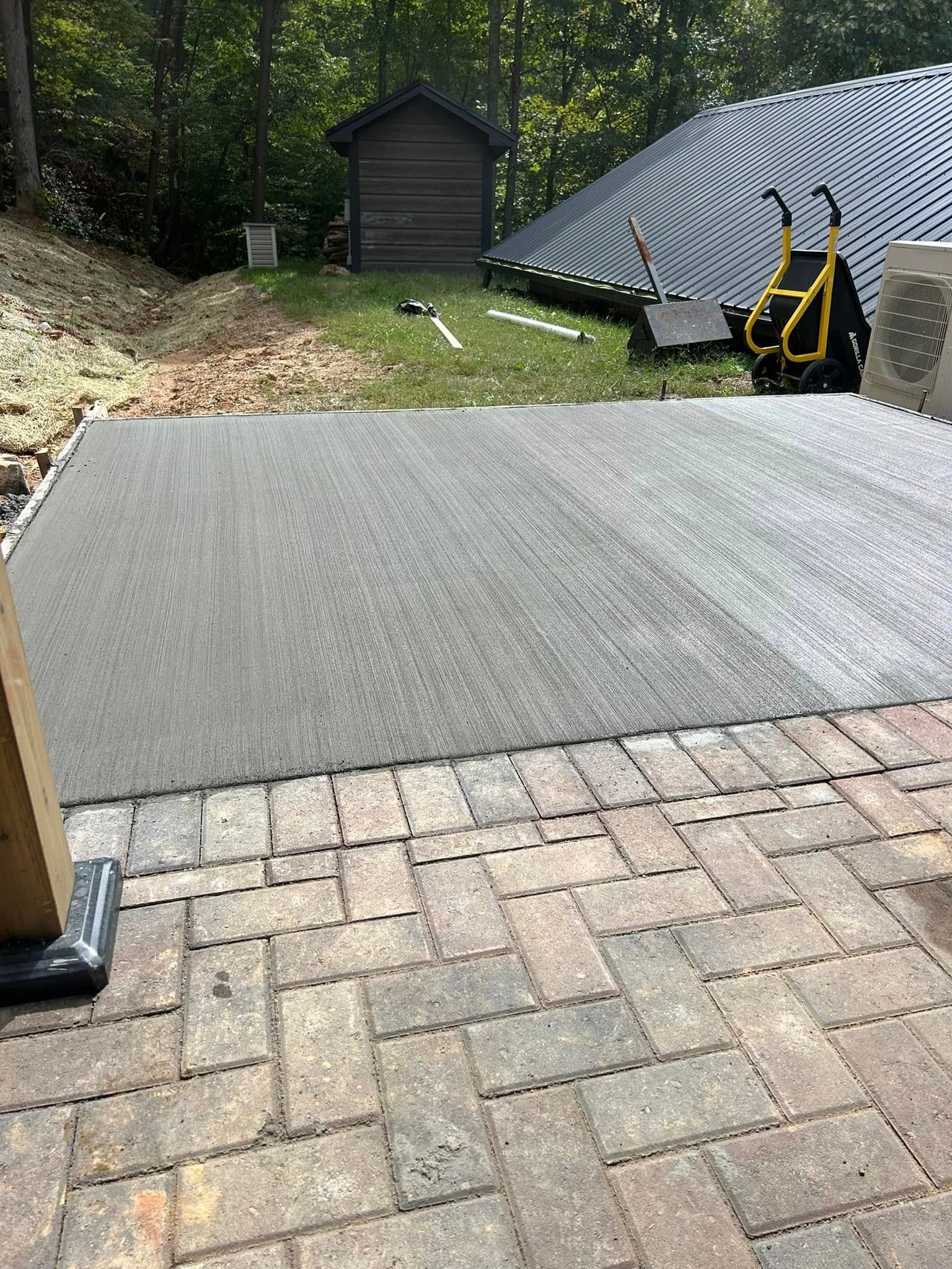 Newly poured concrete patio next to brick pavers, with a shed and trees in the background.