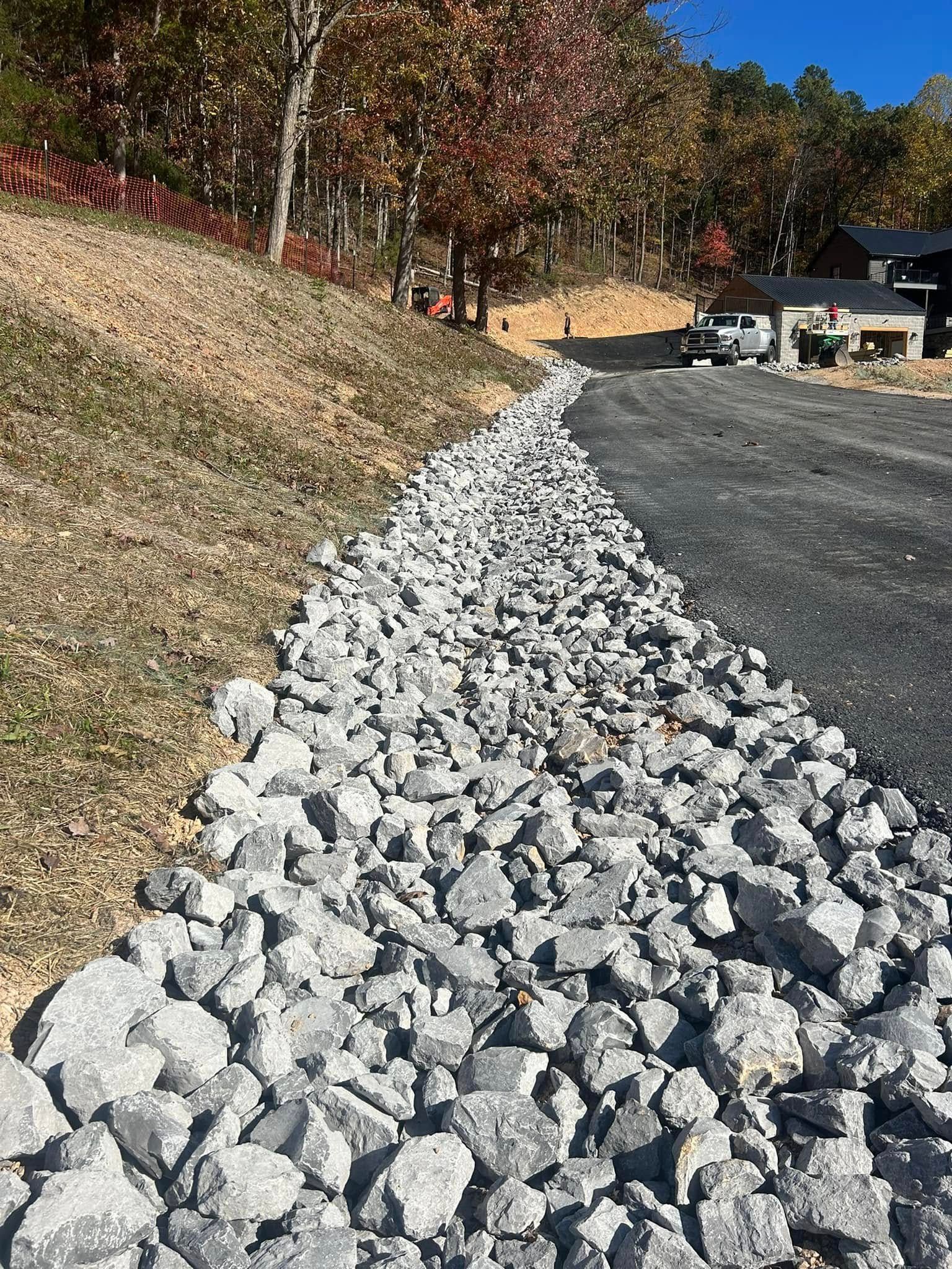 Rocks lining a driveway, next to a grassy slope with trees in the background.