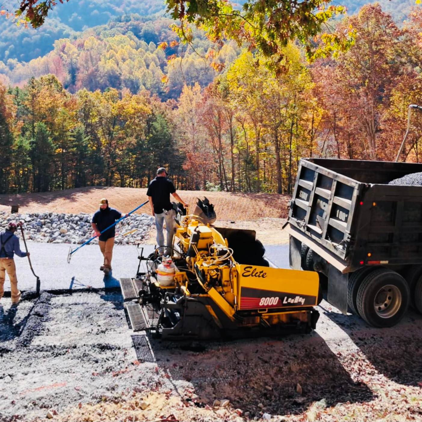 Road paving in progress; workers, asphalt spreader, dump truck, autumn forest background.