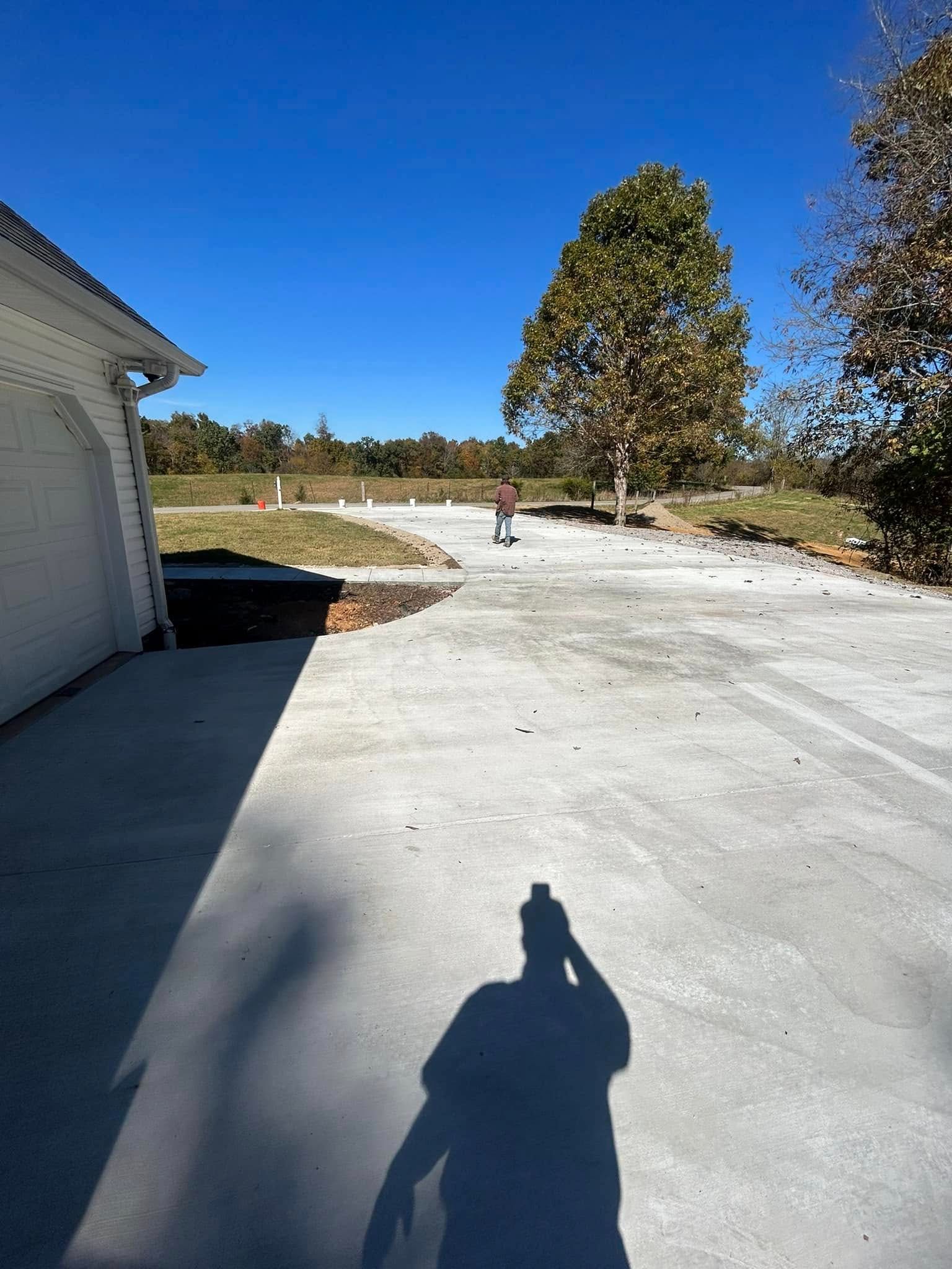 Shadow of a person on a concrete driveway, next to a garage and a tree with fall foliage; sunny day.
