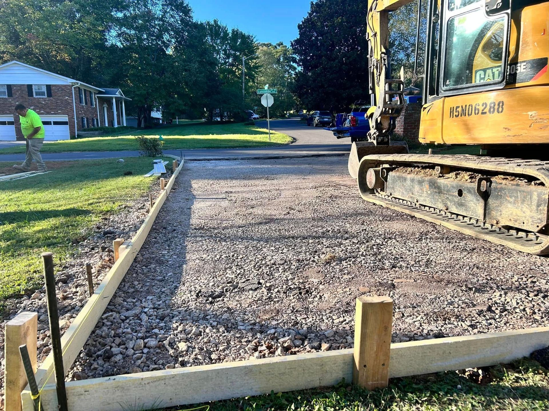 Construction site: Excavator beside a gravel-filled form for a driveway. Worker in background.