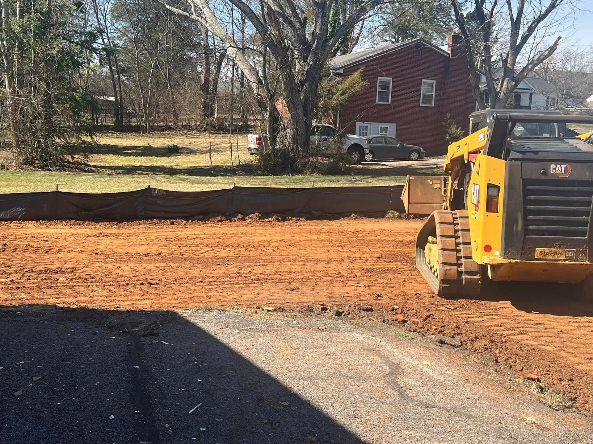 Yellow skid steer grading dirt in front of a red brick house; sunny day.