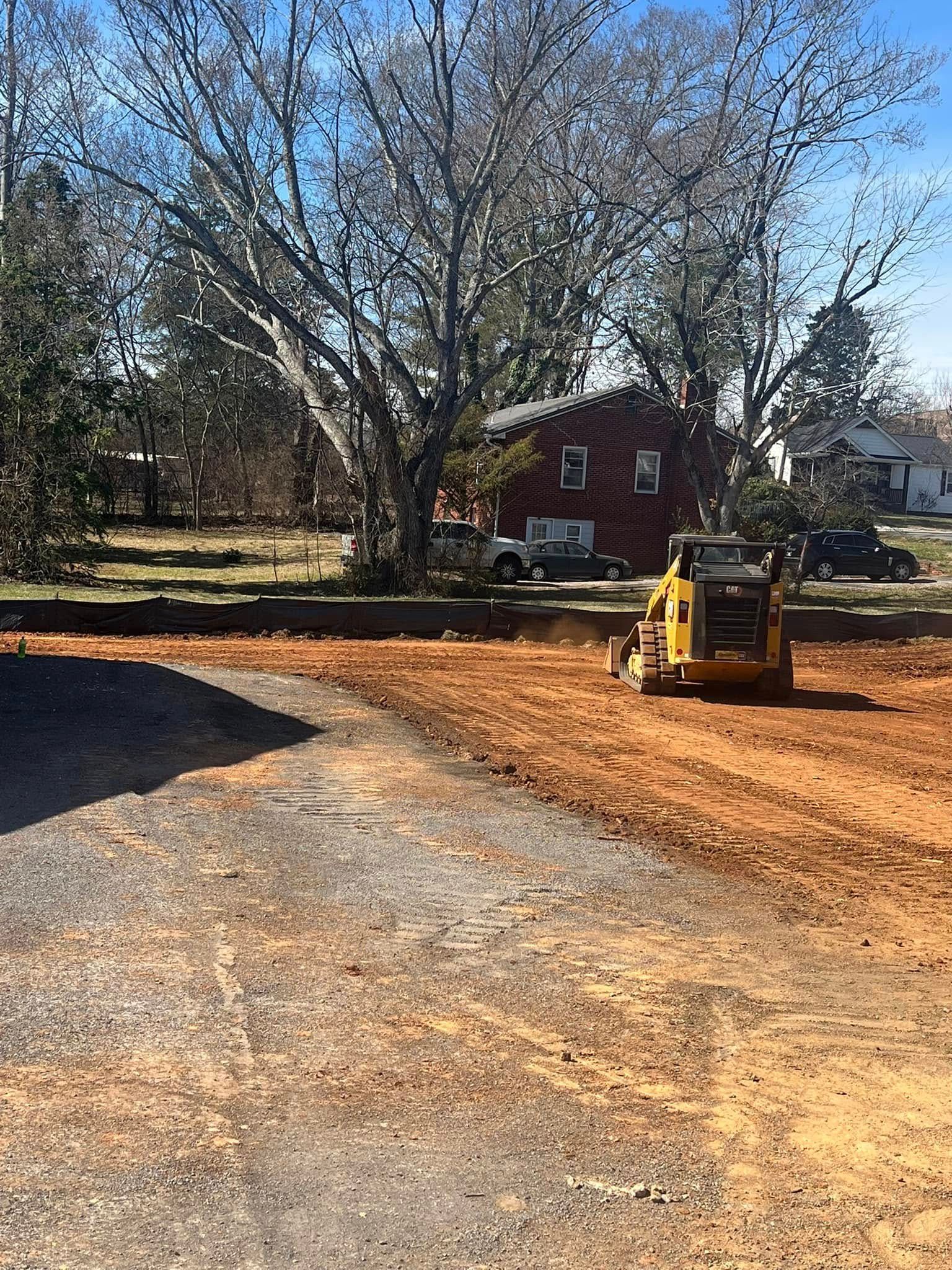 A yellow skid steer compacts red dirt on a construction site. A red brick house is in the background.