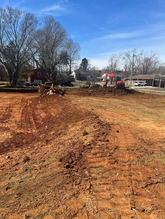 Dirt field with red excavator working, trees and houses in the background on a sunny day.