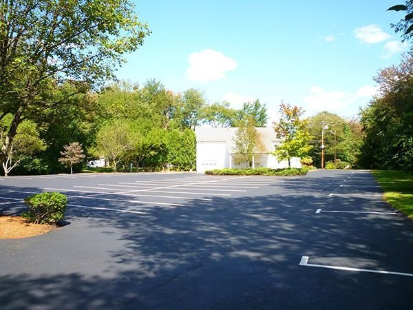 Empty asphalt parking lot in front of a white building, surrounded by green trees under a blue sky.