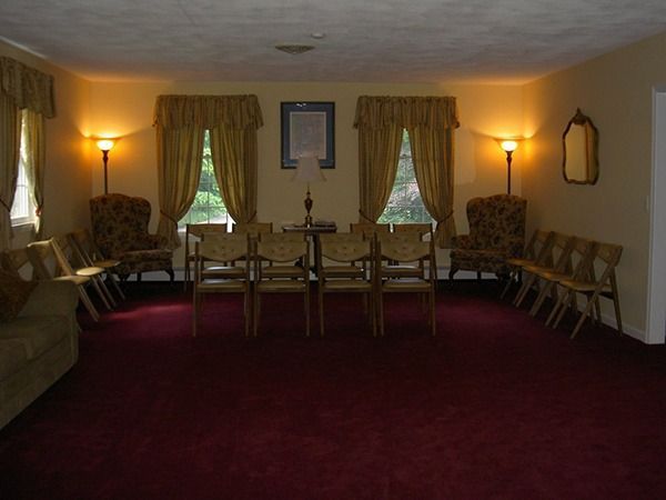 Funeral home viewing room, red carpet, chairs arranged in rows, two windows with curtains, side lamps.