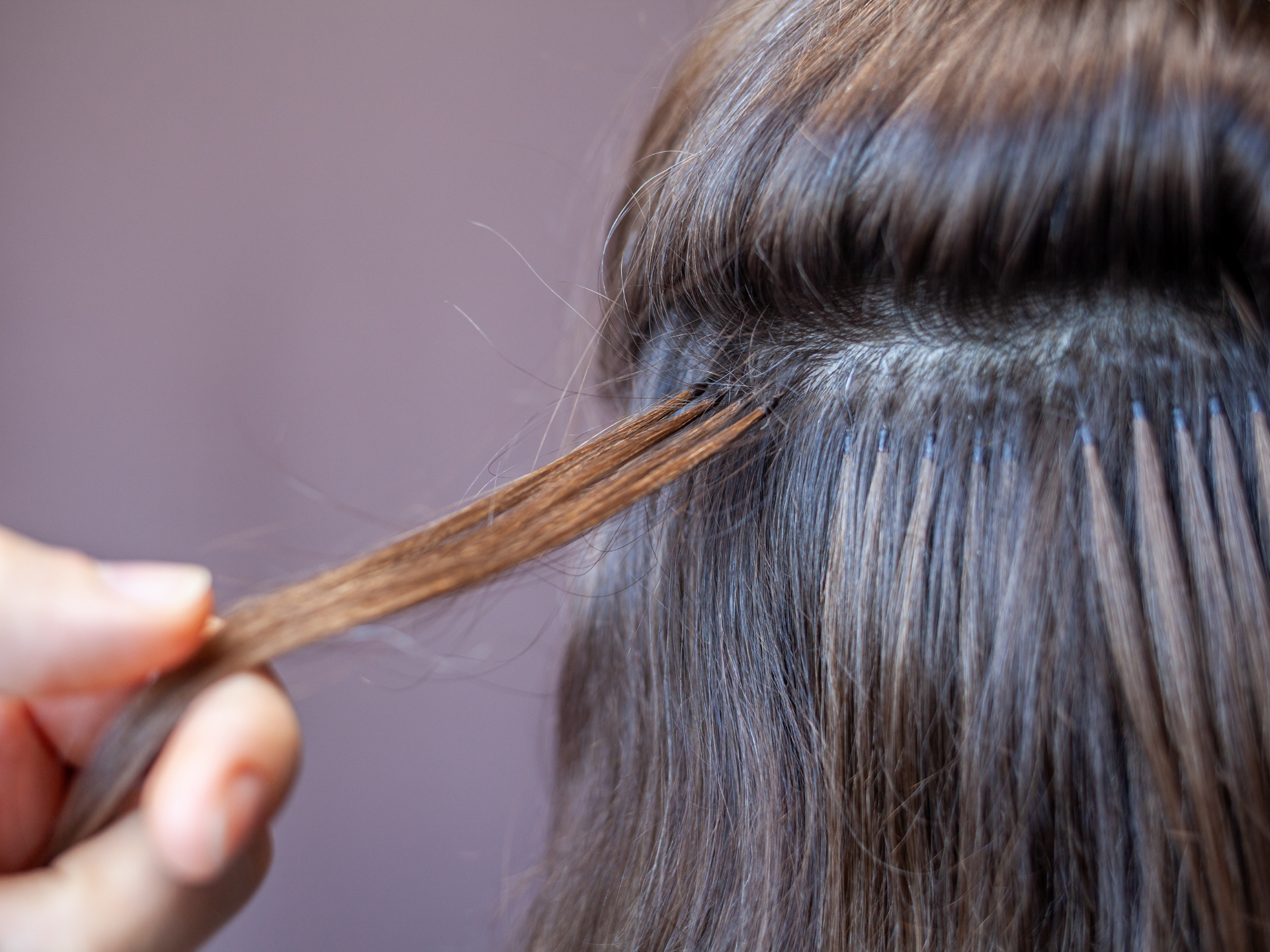 Hand holding a strand of brown hair, showing hair extensions attached to the scalp with small beads.