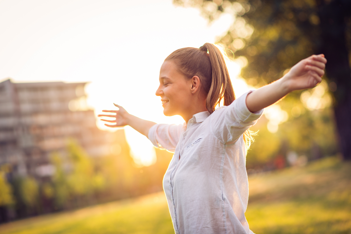 Woman with arms outstretched, smiling in sunlight outdoors.