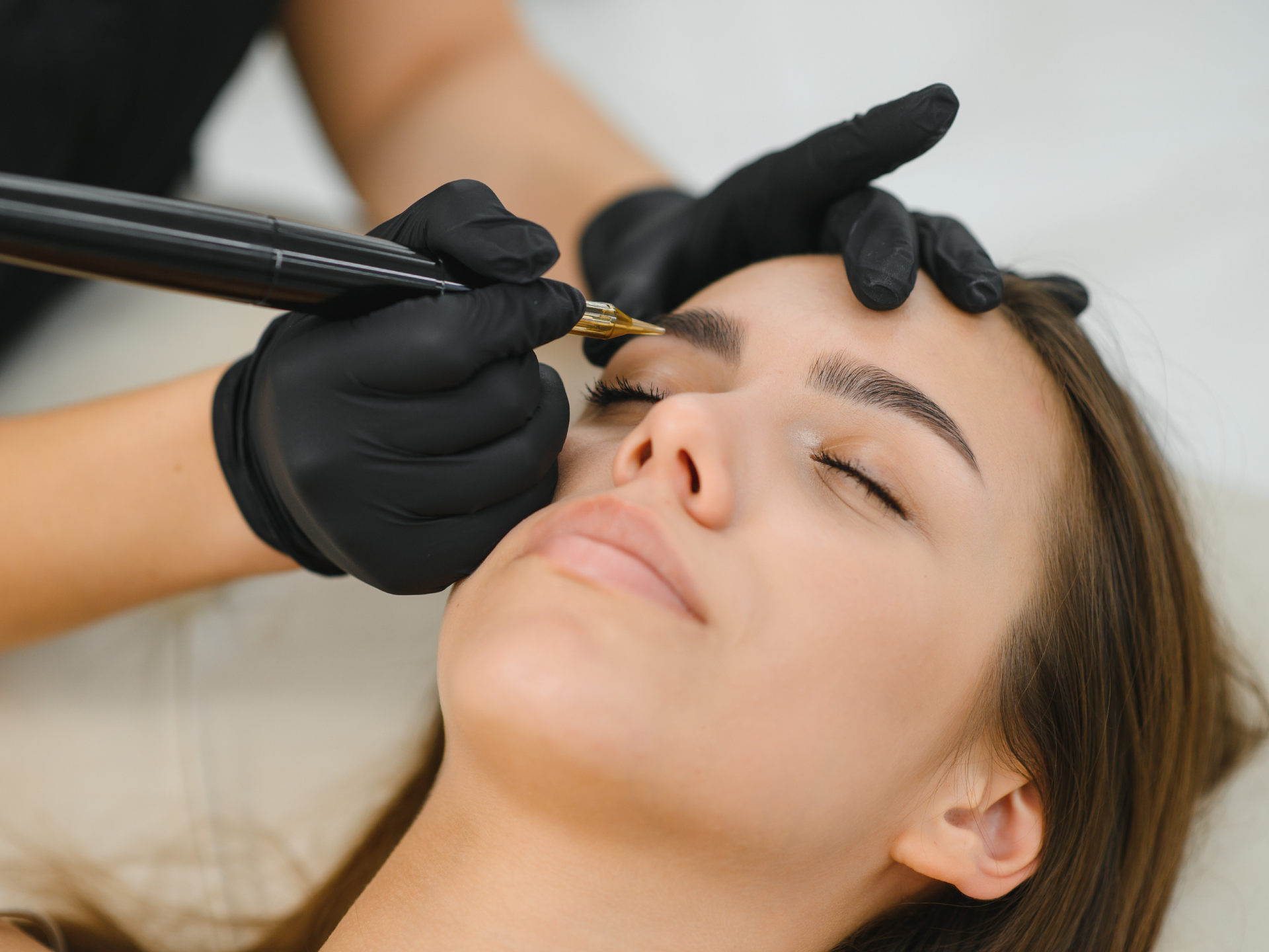 Person receiving eyebrow microblading with black-gloved hands holding a tool near her closed eye.