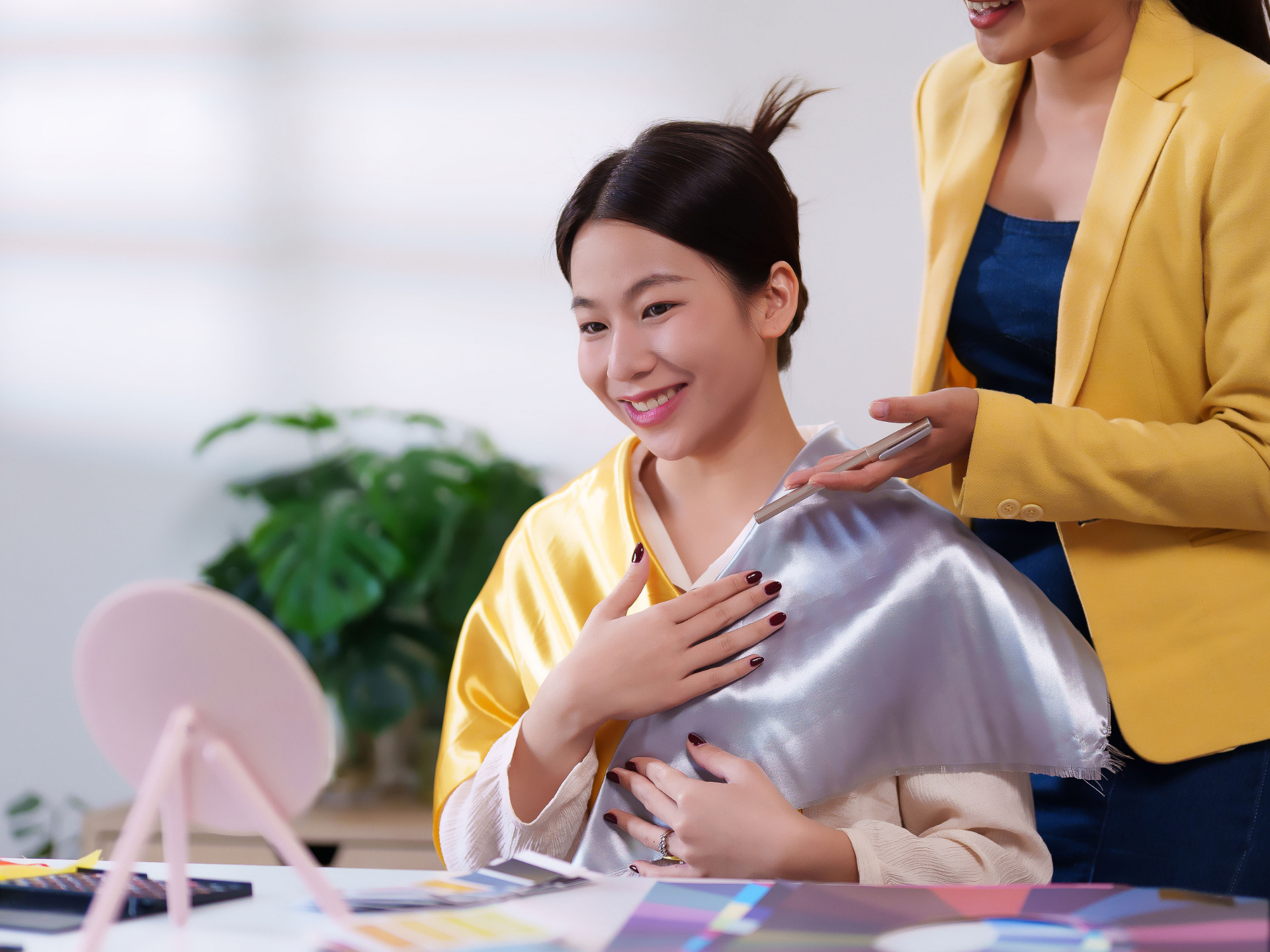 Woman smiles, holding silver fabric, being color-consulted by a person in a yellow jacket. Indoors,