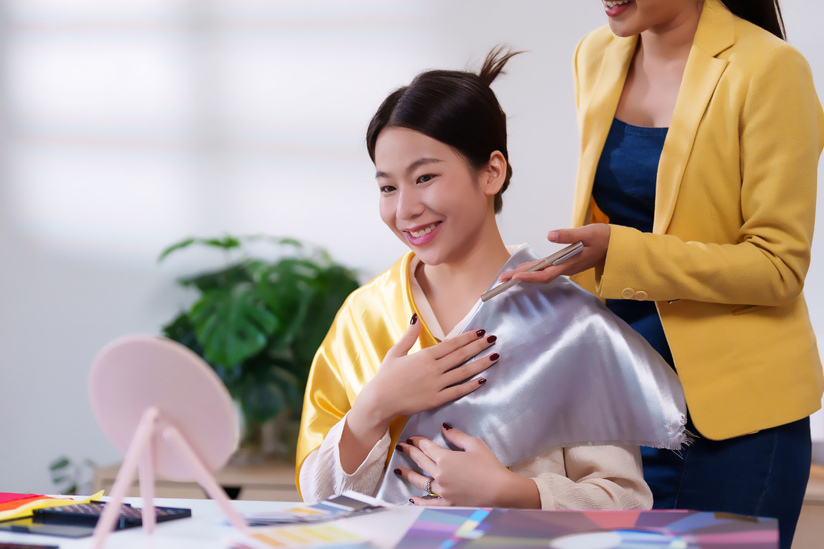 Woman smiling during a color analysis session. A stylist holds a silver and yellow fabric up to her, indoors.
