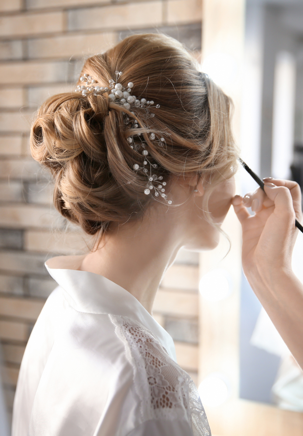 Woman's updo hairstyle with pearl hairpiece, getting makeup applied. Indoors, near a brick wall.