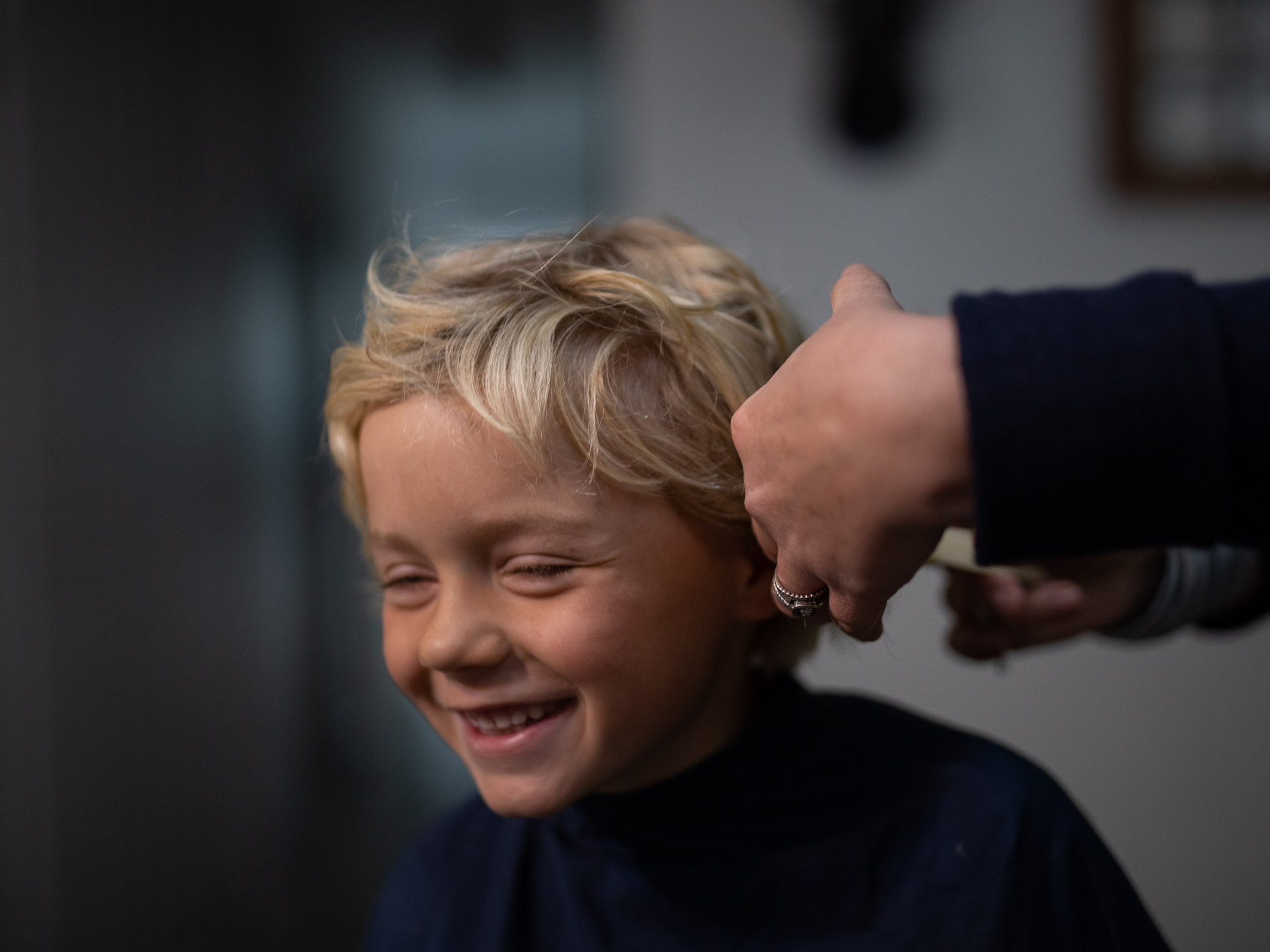 Boy smiles while getting a haircut; hand holds scissors near his ear.