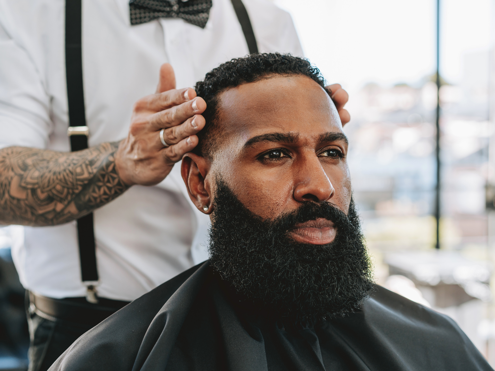 Barber styling a man's hair, hands over his head. Man with full beard, in barber chair.