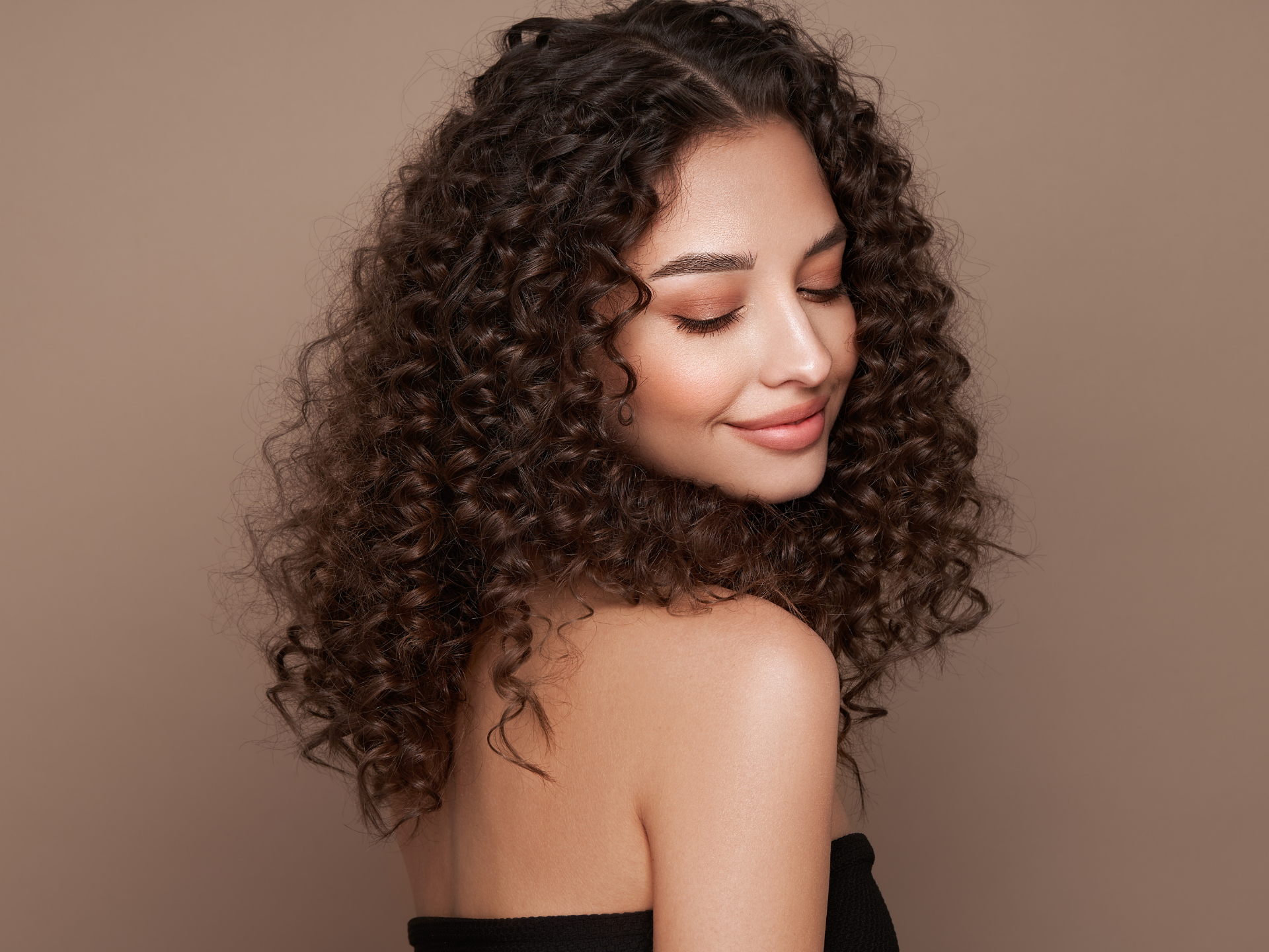 Woman with dark, curly hair, smiling with eyes closed, shoulders bare, against a light brown background.