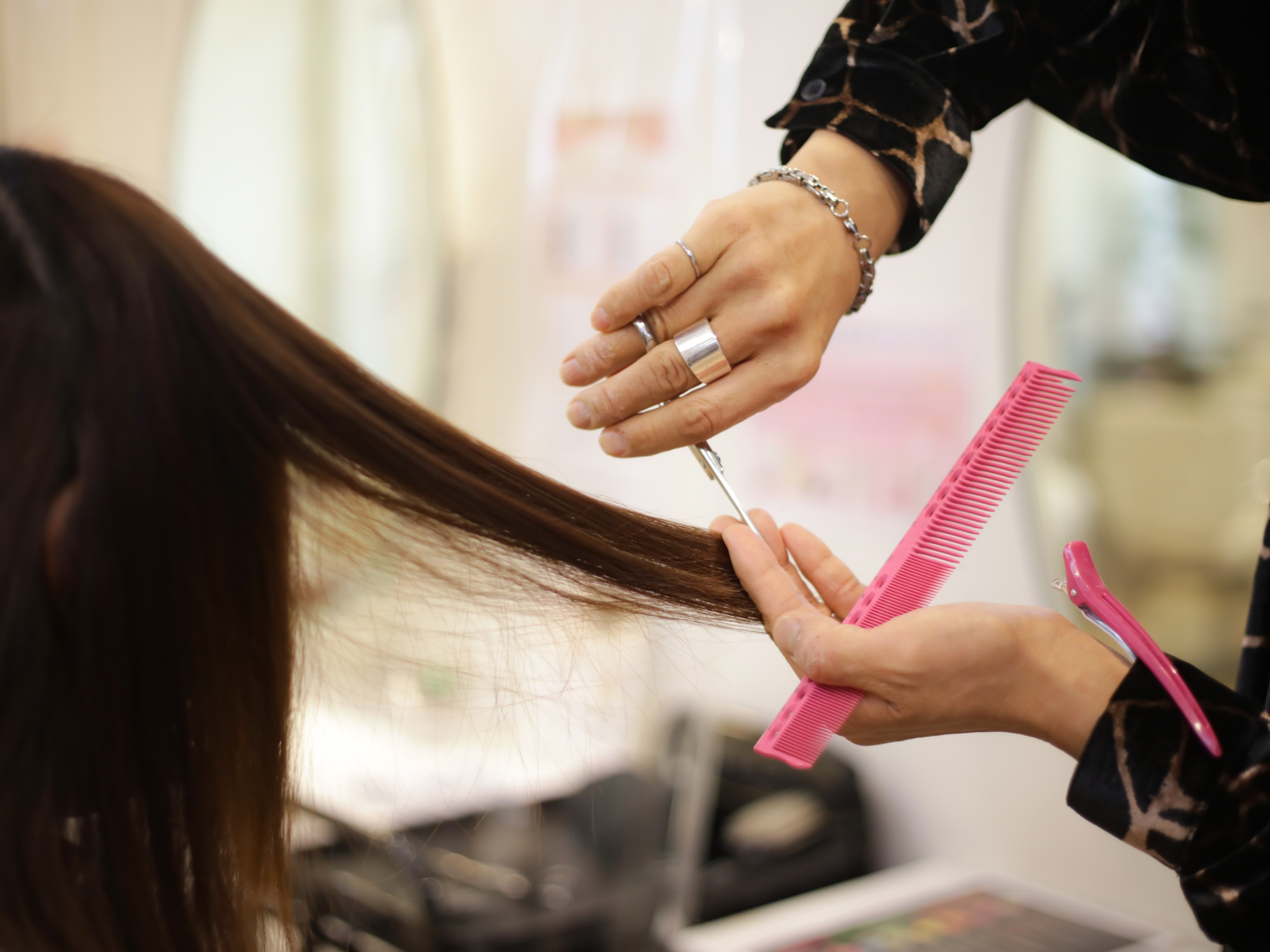 Hair stylist cutting a client's long, dark hair with scissors and a pink comb in a salon.